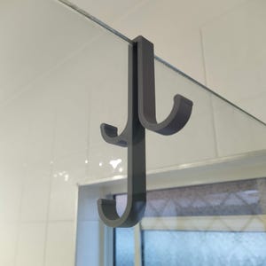 Peut inclure: Un crochet de porte gris foncé avec trois crochets incurvés. Le crochet est conçu pour être accroché sur une porte de douche en verre. Le fond montre un mur carrelé blanc et une fenêtre.