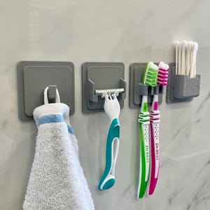 Peut inclure: Ensemble d'organisateurs de salle de bain adhésifs gris. Comprend un crochet avec une serviette blanche et bleue, un support de rasoir avec un rasoir turquoise et blanc, des porte-brosses à dents avec des brosses vertes et roses et un porte-coton-tige.