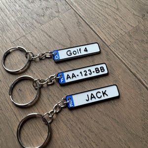 Peut inclure: Trois porte-clés avec des étiquettes rectangulaires blanches et des bordures noires. Les étiquettes affichent le texte "Golf 4", "AA-123-BB" et "JACK". Chaque porte-clés a un anneau et une chaîne argentés, sur fond de bois.