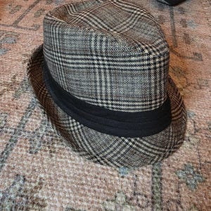 May include: A fedora hat with a plaid pattern in shades of brown, tan, and black. The hat features a black band around the crown. The hat is a classic style, suitable for various occasions.