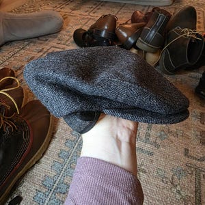 以下が含まれることがあります： 丸みを帯びたクラウンと小さく硬いブリムが付いた、グレーのツイードニュースボーイキャップ。キャップはヘリンボーン柄で、濃い無地のブリムが付いています。靴と犬用ベッドを背景に、キャップが掲げられています。