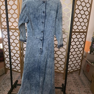 May include: A long-sleeved, acid-wash denim dress with a button-up front and a flared skirt. The dress is hanging on a black clothing rack. The dress has a round neckline and long sleeves with buttoned cuffs.