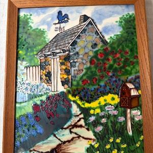 May include: A framed ceramic tile featuring a colorful garden scene. The artwork depicts a stone cottage with a weathervane, surrounded by vibrant flowers and a pathway. The frame is made of wood.