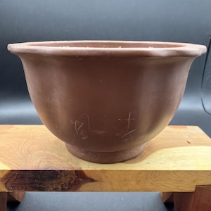 May include: A matte brown clay bowl with a wide, flared rim and a circular base. The exterior surface features two distinct, lightly etched calligraphic characters in a light brown tone, positioned centrally on the front of the rounded vessel.