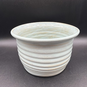 May include: A light blue ceramic planter with a wide, circular opening and a series of horizontal, raised ridges encircling the exterior. The glaze has a subtle, glossy finish with faint variations in color tone across the curved surface.