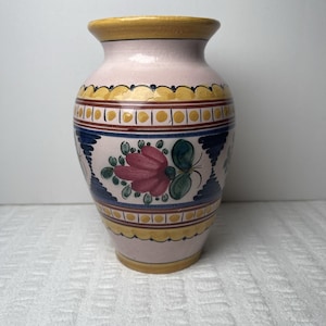 May include: A ceramic vase with a light pink base color, featuring a hand-painted design of dark blue vertical patterns and pink flowers with green leaves. The rim and base are accented with yellow scalloped borders and brown lines.