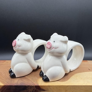 May include: A pair of white ceramic napkin rings shaped like sitting pigs. Each pig features a glossy white finish, a small pink snout, black painted eyes, and black hooves. The circular opening for the napkin is integrated into the pig's body.