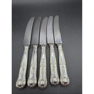 May include: A set of five silver-toned dinner knives featuring smooth, curved blades and ornate, embossed handles with a shell-like motif at the base. The metal surfaces exhibit a polished finish with intricate decorative detailing.