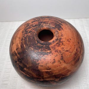 May include: A round, terracotta-colored ceramic vase with a dark brown interior and a circular opening at the top. The exterior features a mottled pattern of brown and orange hues, creating a rustic aesthetic.