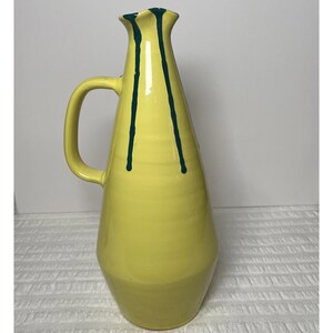 May include: A tall, yellow ceramic pitcher with a glossy finish and a single handle. The vessel features a tapered neck and a wider base, decorated with two distinct dark green vertical drip patterns that extend from the rim down the body.