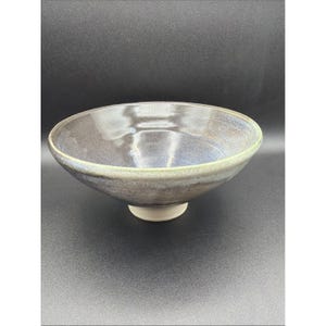 May include: A wide, shallow ceramic bowl with a glossy, dark blue and purple interior glaze that transitions to a lighter, pale green rim. The exterior features a smooth, dark finish, and the bowl rests on a small, unglazed, light-colored circular base.