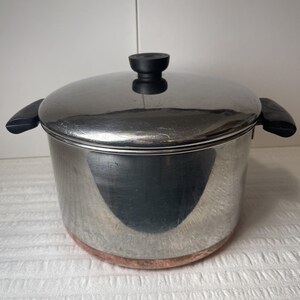 May include: A stainless steel stockpot with a polished finish, featuring a matching lid with a black knob and two black side handles. The base of the pot is constructed from copper, providing a distinct contrast to the silver-toned body.