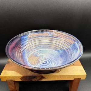 May include: A round ceramic bowl featuring a swirling glaze pattern in shades of deep blue, violet, and earthy tan. The interior surface displays concentric circular ridges that spiral toward the center, creating a textured appearance.