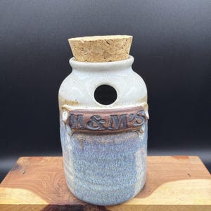 May include: A cylindrical ceramic jar with a speckled off-white top and a blue-toned base. The jar features a circular opening near the top and a raised brown rectangular plaque with the embossed text M&M'S. It is sealed with a cork stopper.