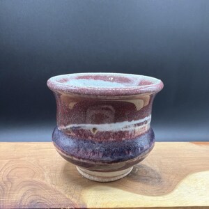 May include: A small, rounded ceramic cup featuring a variegated glaze in shades of deep purple, maroon, and white. The vessel has a wide, flared rim, a curved body that tapers toward the base, and a smooth, glossy finish with visible color blending.