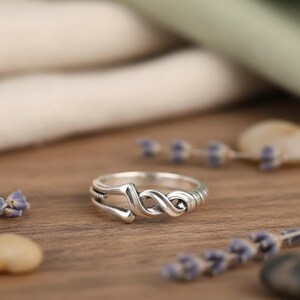 May include: A silver-colored ring with an infinity symbol design. The ring features a twisted, figure-eight pattern in the center, with a smooth band and decorative detailing. The ring is displayed on a wooden surface with small purple flowers.