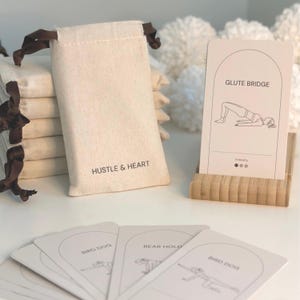 May include: A set of off-white exercise cards featuring line drawings of fitness poses like Glute Bridge, Bird Dog, and Bear Hold. The cards are displayed in a bamboo stand and stored in a beige cotton drawstring pouch labelled Hustle & Heart.