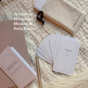 May include: A set of white cards with the word "GROUND" printed on them, alongside a pen, a beige pouch with the text "HUSTLE & HEART", and a notebook. The text "36 Cards for Movement Mindset & Daily Resets" is also visible.