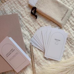 Può includere: Un set di carte bianche con la parte superiore arrotondata e la parola "GROUND" stampata, insieme a una penna e un sacchetto beige con le parole "Hustle & Heart". Sono visibili anche un taccuino rosa e un mazzo di carte con il testo "Hustle & Heart Movement Cards".
