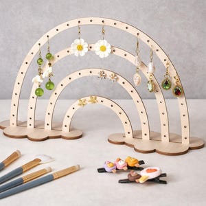 Puede incluir: Un expositor de pendientes de madera en forma de arcoíris, con múltiples niveles, que muestra varios pendientes. El expositor es de color marrón claro con bases en forma de nube. Varios pares de pendientes, incluyendo diseños florales y de gemas, cuelgan de los agujeros. Brochas de maquillaje y pinzas para el pelo en primer plano.