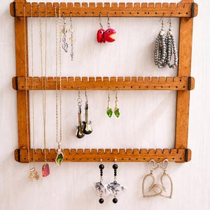 May include: A wooden jewelry organizer with three horizontal bars, displaying various earrings and necklaces. The earrings include red fish, green gems, and beaded styles. Necklaces with pendants hang alongside. The organizer is made of light brown wood.