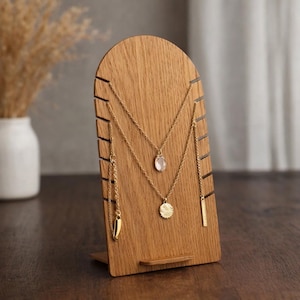 May include: A wooden jewellery display stand with an arched top. The stand holds several gold necklaces with various pendants, including a teardrop, a circular pendant, and a bar. The wood has a natural grain and a warm, light brown colour.