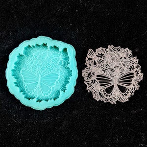 Può includere: Uno stampo in silicone turchese con un designo dettagliato di farfalle e fiori. Lo stampo è accanto a un ritaglio in metallo argentato dello stesso design.