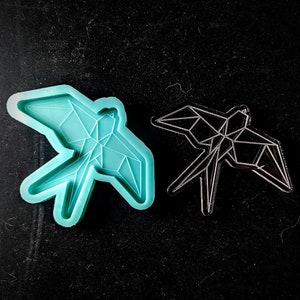 Puede incluir: Un molde de silicona azul turquesa con un diseño de pájaro geométrico. El molde está al lado de un recorte de metal negro del mismo diseño de pájaro.