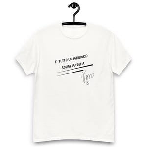 Può includere: Una t-shirt bianca a maniche corte con il testo nero E' TUTTO UN EQUILIBRIO SOPRA LA FOLLIA, seguito da due linee orizzontali e una firma stilizzata. Il capo è mostrato su una gruccia nera su uno sfondo bianco uniforme.