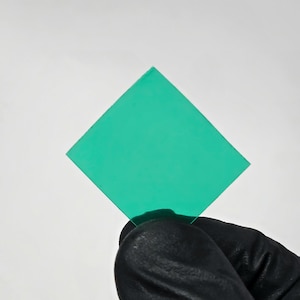 May include: A square, transparent, teal-colored object is held against a white background. The object is a perfect square with sharp corners. The hand holding the object is covered by a black glove.