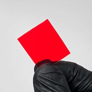 May include: A vibrant red square held by a gloved hand against a white background. The square appears to be made of a translucent material, allowing light to pass through. The edges are clean and well-defined.