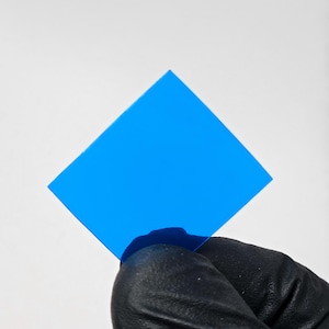 Puede incluir: Un cuadrado azul cian brillante de material transparente se sostiene sobre un fondo blanco. El objeto es de forma sólida y plana, con un acabado brillante. Una mano enguantada de negro lo sostiene.