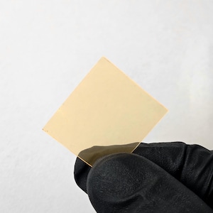 Puede incluir: Una lámina cuadrada, translúcida, de color ámbar, de vidrio o plástico, es sostenida por una mano enguantada sobre un fondo blanco. La lámina es fina y parece ligeramente reflectante.