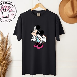 May include: Black t-shirt featuring a cartoon Mickey Mouse dressed as a doctor, complete with a stethoscope and clipboard. The design includes pink shoes and a light blue shirt. The shirt is hanging on a wooden hanger.
