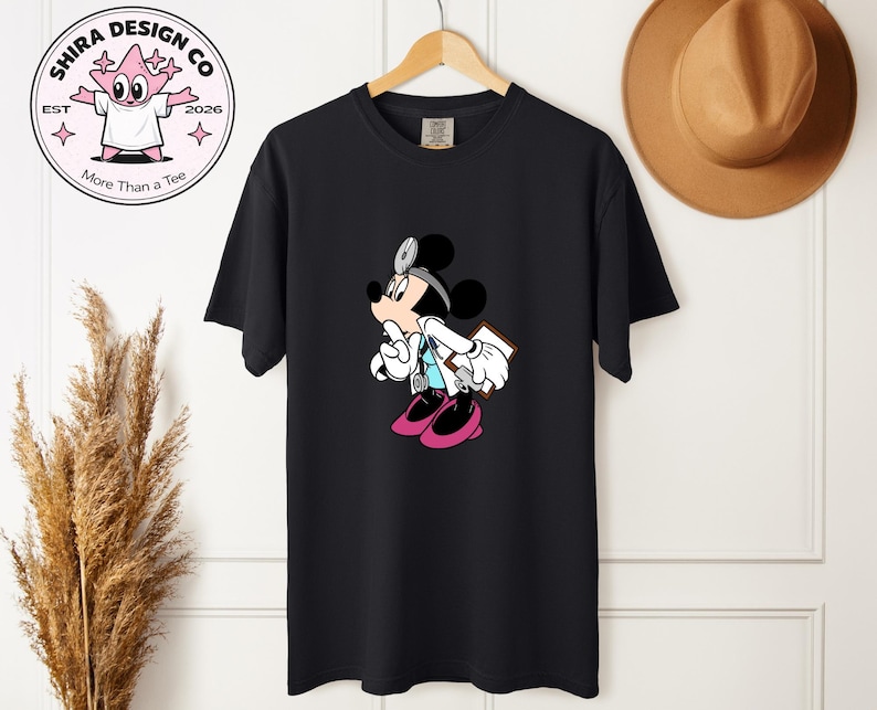 May include: Black t-shirt featuring a cartoon Mickey Mouse dressed as a doctor, complete with a stethoscope and clipboard. The design includes pink shoes and a light blue shirt. The shirt is hanging on a wooden hanger.