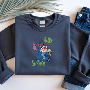 May include: A dark grey sweatshirt with a cartoon character wearing sunglasses and eating ice cream, with the words "hello summer" in green script. The sweatshirt is paired with blue jeans.
