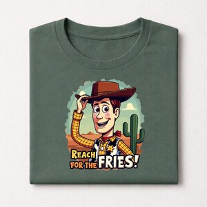 Op de afbeelding: Groen t-shirt met een cartoon illustratie van een cowboy met een bruine hoed, geel geruit overhemd en rode halsdoek. De tekst "REACH FOR THE FRIES!" staat onder de cowboy.
