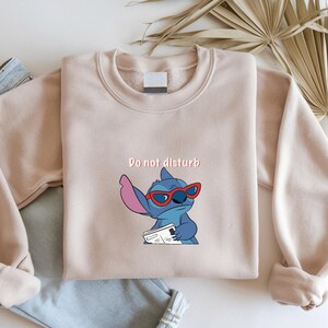 Peut inclure: Un sweat-shirt beige avec l'inscription "Do not disturb" au-dessus d'un personnage de dessin anim&eacute; portant des lunettes de soleil rouges. Le personnage bleu avec de grandes oreilles tient un papier. Le sweat-shirt est pli&eacute; sur un jean bleu.