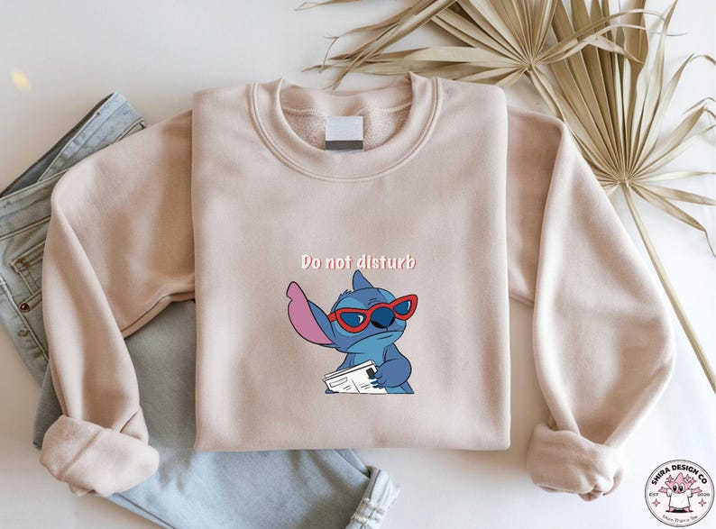 Peut inclure: Un sweat-shirt beige avec l'inscription "Do not disturb" au-dessus d'un personnage de dessin anim&eacute; portant des lunettes de soleil rouges. Le personnage bleu avec de grandes oreilles tient un papier. Le sweat-shirt est pli&eacute; sur un jean bleu.