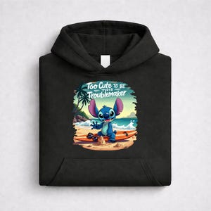 Op de afbeelding: Zwarte hoodie met een afbeelding van een blauw stripfiguur op een strand met een surfplank. De afbeelding bevat de tekst "Too Cute to Be This Troublemaker."