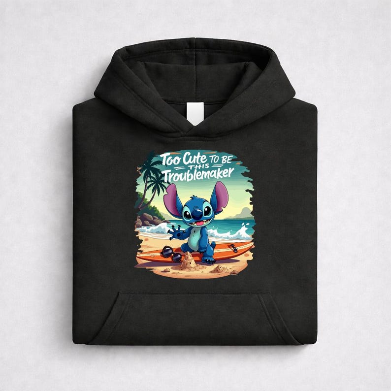 Op de afbeelding: Zwarte hoodie met een afbeelding van een blauw stripfiguur op een strand met een surfplank. De afbeelding bevat de tekst "Too Cute to Be This Troublemaker."
