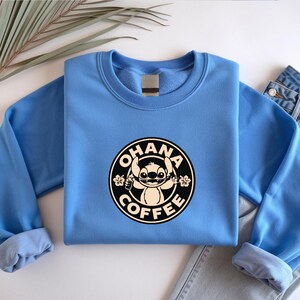 May include: A light blue sweatshirt featuring a circular graphic with the text "OHANA COFFEE" and a cartoon character. The sweatshirt is paired with a pair of blue jeans. The design is in black and white.