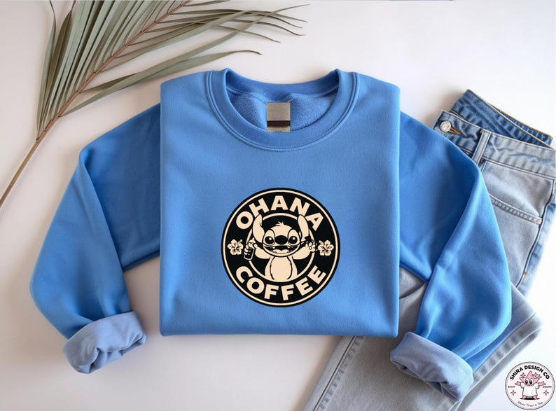 May include: A light blue sweatshirt featuring a circular graphic with the text "OHANA COFFEE" and a cartoon character. The sweatshirt is paired with a pair of blue jeans. The design is in black and white.