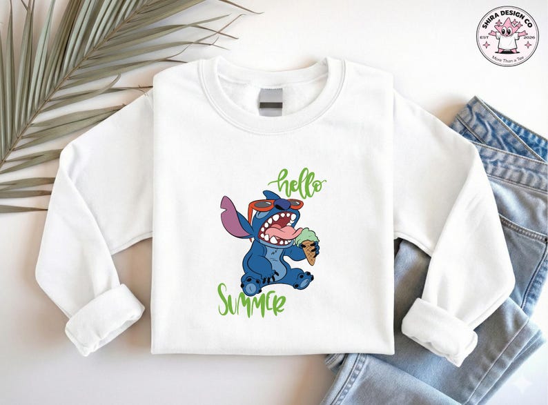 May include: White sweatshirt with a cartoon Stitch design, wearing sunglasses and eating ice cream. Green script reads "hello" and "summer". The sweatshirt is shown with blue denim jeans.