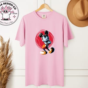 May include: A pale pink t-shirt with a cartoon image of Mickey Mouse in a dynamic pose, set against a red circle. The shirt also displays the text "Shira Design Co" and "More Than a Tee" with a cartoon character logo.