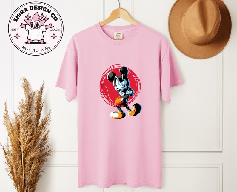 May include: A pale pink t-shirt with a cartoon image of Mickey Mouse in a dynamic pose, set against a red circle. The shirt also displays the text "Shira Design Co" and "More Than a Tee" with a cartoon character logo.