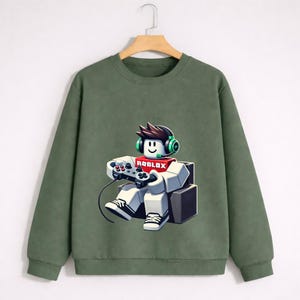 Pu&ograve; includere: Felpa verde oliva con grafica di un personaggio Roblox con cuffie e controller di gioco. Il personaggio &egrave; seduto e la parola "ROBLOX" &egrave; sul petto. La felpa &egrave; appesa a una gruccia di legno.