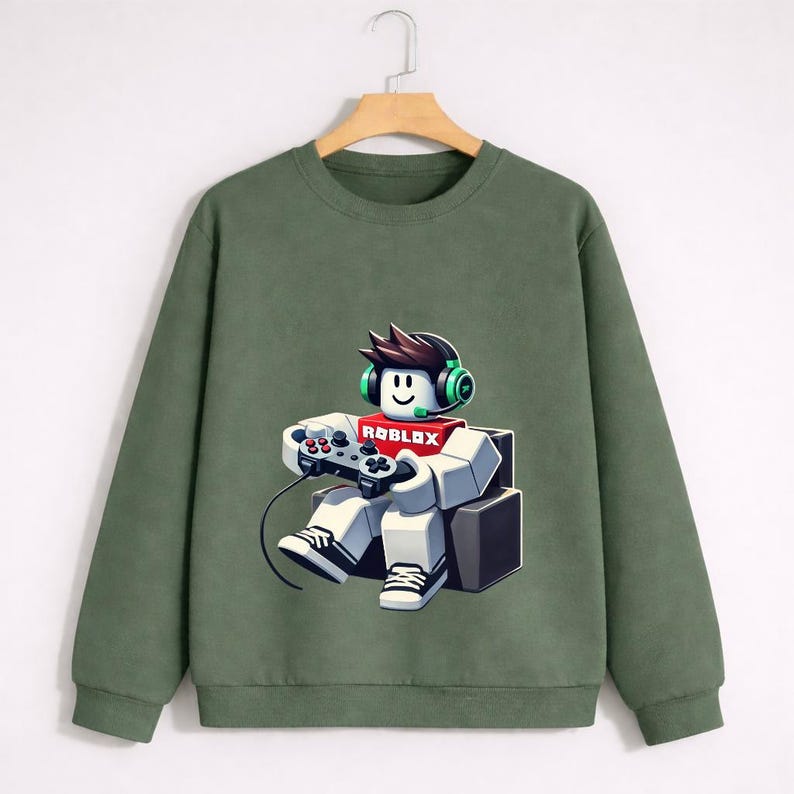 Pu&ograve; includere: Felpa verde oliva con grafica di un personaggio Roblox con cuffie e controller di gioco. Il personaggio &egrave; seduto e la parola "ROBLOX" &egrave; sul petto. La felpa &egrave; appesa a una gruccia di legno.