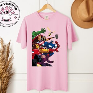 Może przedstawiać: Jasnor&oacute;żowa koszulka z kolorową grafiką przedstawiającą superbohater&oacute;w w akcji. Koszulka wisi na drewnianym wieszaku. Logo marki "Shira Design Co" jest widoczne w lewym g&oacute;rnym rogu.