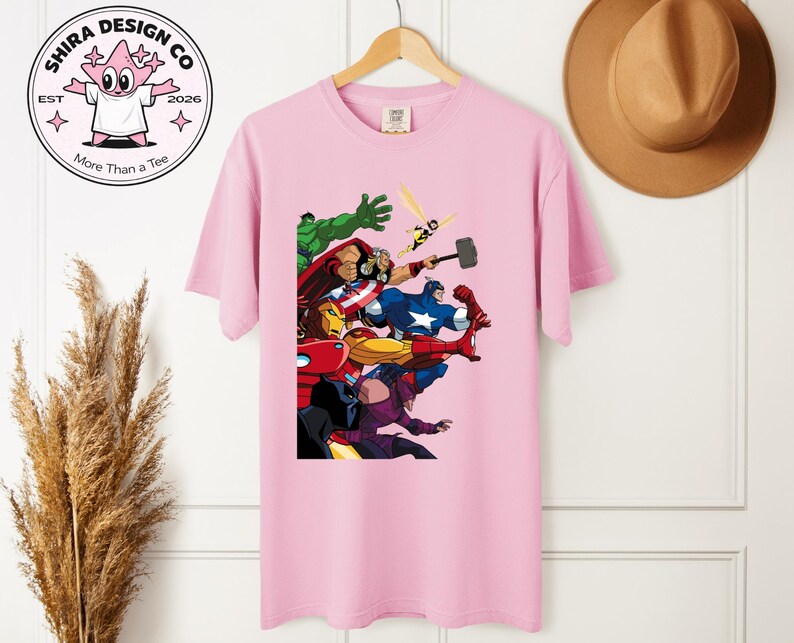 Może przedstawiać: Jasnor&oacute;żowa koszulka z kolorową grafiką przedstawiającą superbohater&oacute;w w akcji. Koszulka wisi na drewnianym wieszaku. Logo marki "Shira Design Co" jest widoczne w lewym g&oacute;rnym rogu.